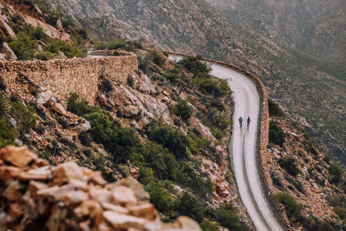 Two riders on a gravel road winding through a rocky mountain pass